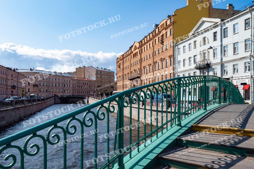 Эта фотография представляет Сенной мост в Санкт-Петербурге в ясный день. Мост выкрашен в характерный зелёный цвет, и на заднем плане видны классические фасады петербургских зданий. Это изображение могло бы хорошо смотреться в статьях о городской жизни Санкт-Петербурга, в туристических буклетах, как часть образовательных материалов по истории города или просто как приятное напоминание о прогулке по Петербургу.
